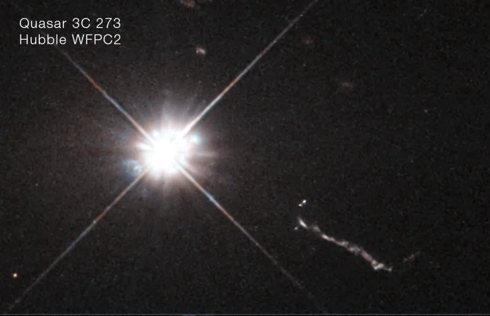 Quasar 3C 273 imaged by Hubble's WFPC2 — a blinding, star-like core with diffraction spikes, throwing a luminous jet toward the lower right.